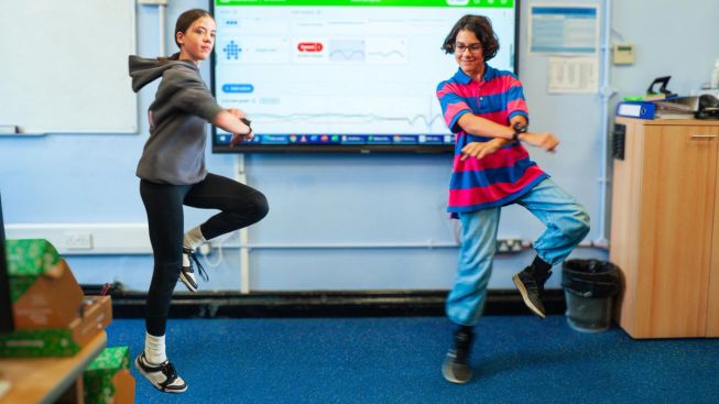 Two students perform a dance routine in a classroom while wearing micro:bits, using movement to interact with code.