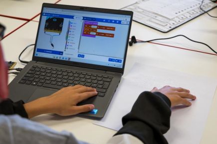 A student feeling a text program embossed in braille, with the corresponding project open in the MakeCode editor on a laptop in front of them.