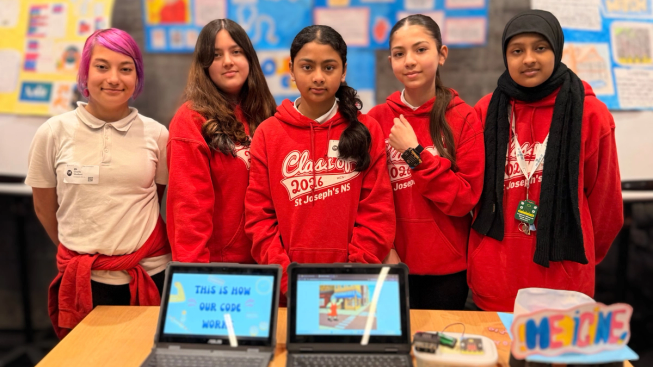 Five learners stand behind a project display at Microsoft Ireland’s Dream Space event. Two laptops show their code alongside a micro:bit prototype and a colourful sign reading “Medicine”. Posters featuring other projects are displayed behind them.