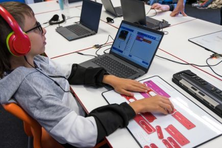 A student feeling a tactile toolbox key showing tactile icons and braille labels for toolbox categories. A laptop in front of them displays the MakeCode editor with a corresponding program open.
