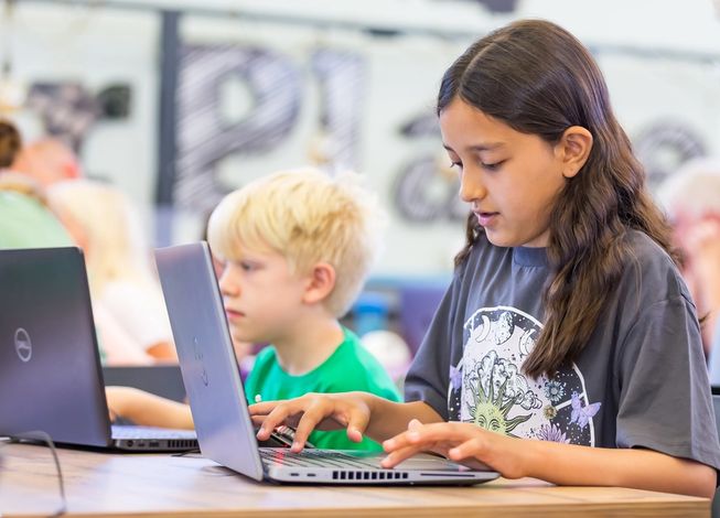 Girl typing on a keyboard in a classroom setting, one boy visible in the background also on a laptop