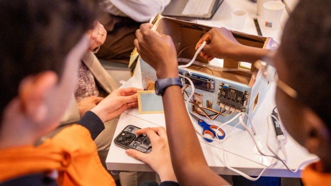 A group of young people work together on a physical computing project using the BBC micro:bit. They collaborate around a cardboard prototype connected with wires, sensors and electronic components as they test and refine their ideas.