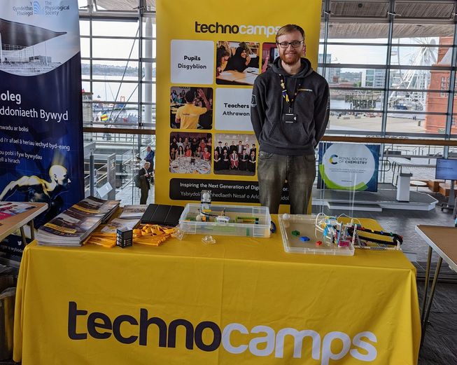 Luke, a Welsh translator, stands behind a bright yellow Technocamps stand