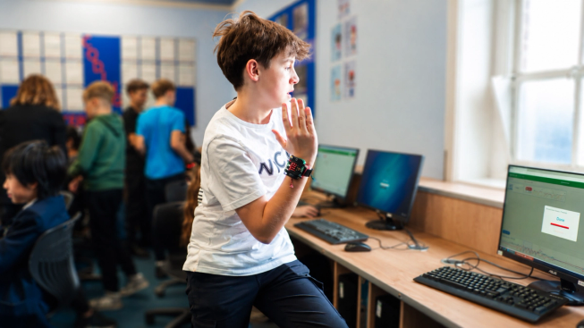 Alt text: A student raises a hand with a BBC micro:bit strapped to their wrist beside a computer displaying movement data graphs, while other students work together in the classroom.
