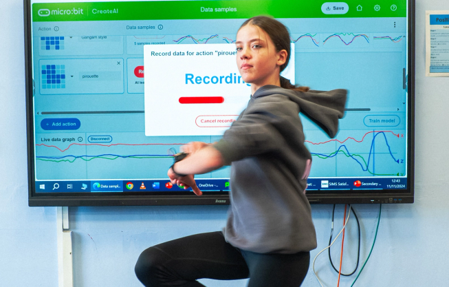 A girl performing a pirouette in front of CreateAI on an electronic whiteboard. She has a micro:bit on her wrist and CreateAI is showing a graph of her movement and a recording progress bar.