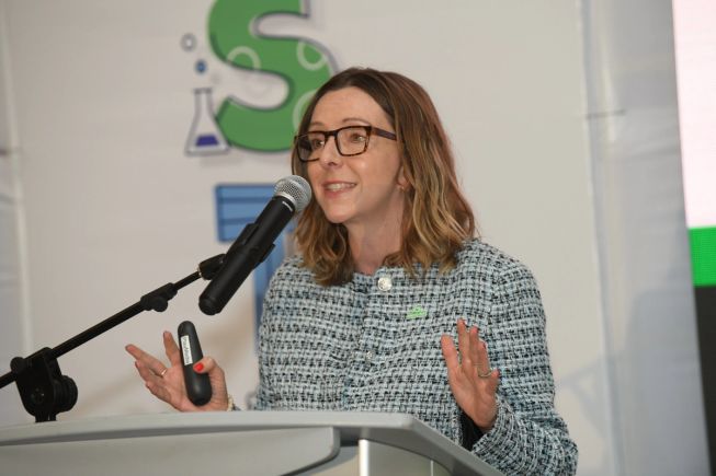 Melanie Washington speaking at a podium with a microphone, wearing a tweed jacket and micro:bit pin, presenting to an audience.