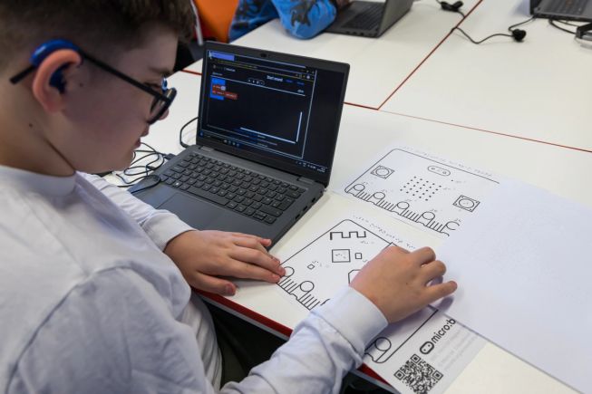 A student uses a laptop running Microsoft MakeCode for micro:bit while exploring tactile diagrams and embossed braille resources on the desk. The tactile resources include raised diagrams of the micro:bit to support accessible coding activities.