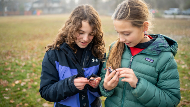 Two children outdoors, each holding and examining a BBC micro:bit connected to a battery pack during a hands-on physical computing activity.