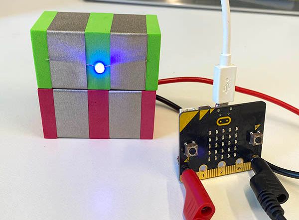 A micro:bit with banana plugs connected to pin 0 and GND, leading to two foam TapeBlocks covered in conductive foil strips. The top TapeBlock holds an LED which is on. The micro:bit is also connected to a USB cable. 