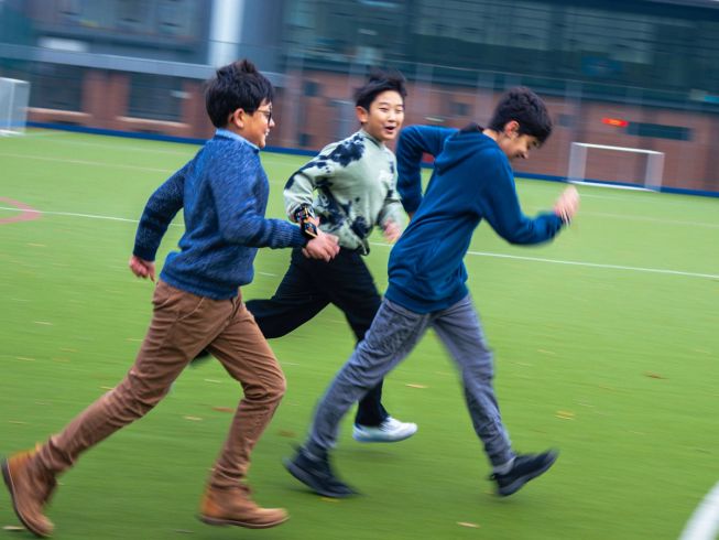 Three boys are running across the field wearing their micro:bits, which are measuring their steps