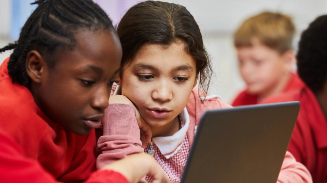 Two primary age girls in school uniform in a classroom working on a laptop.