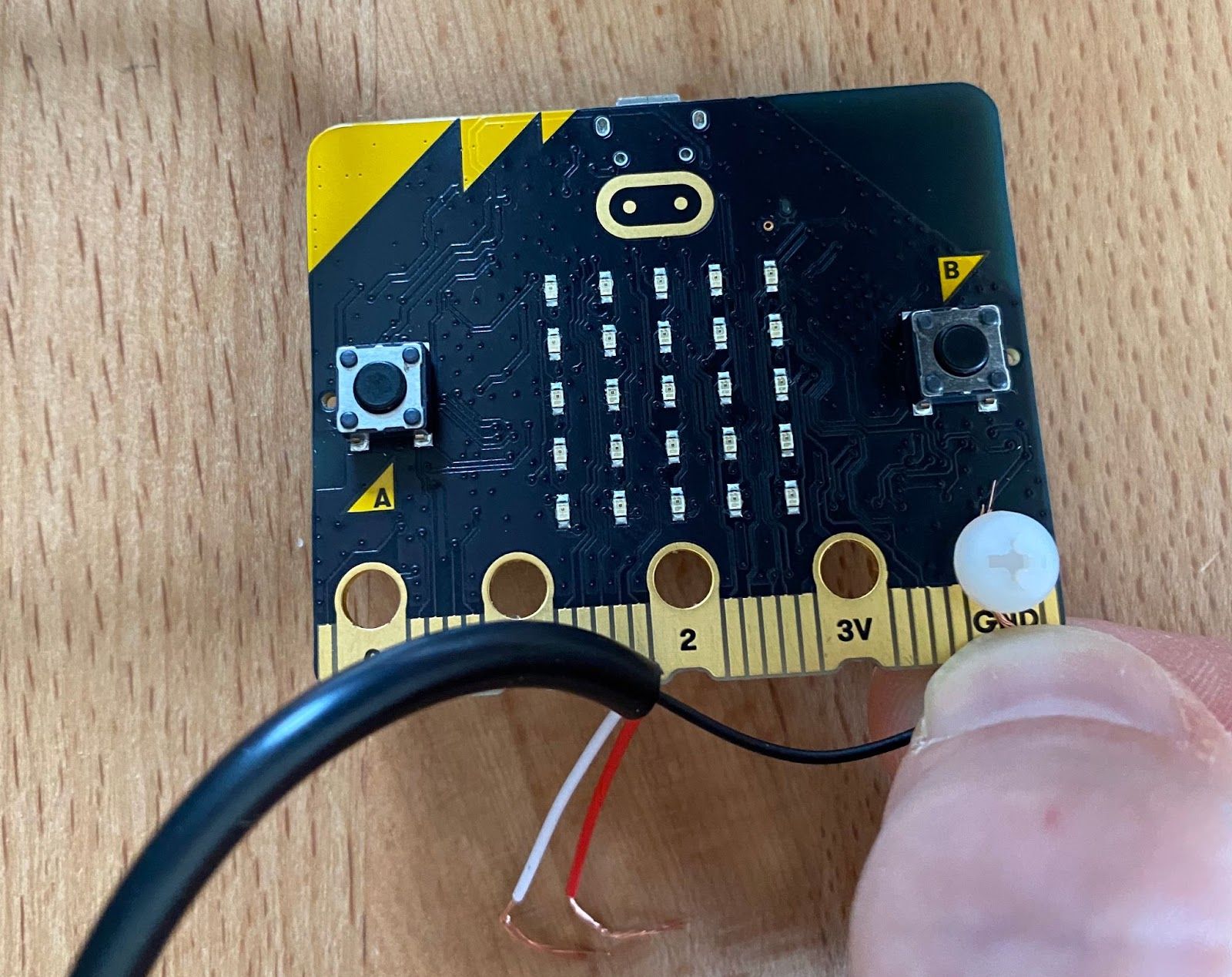 Front view of a micro:bit with a nylon M4 bolt through the GND pin hole, showing someone inserting a wire from a 3-conductor cable with stripped jacket.