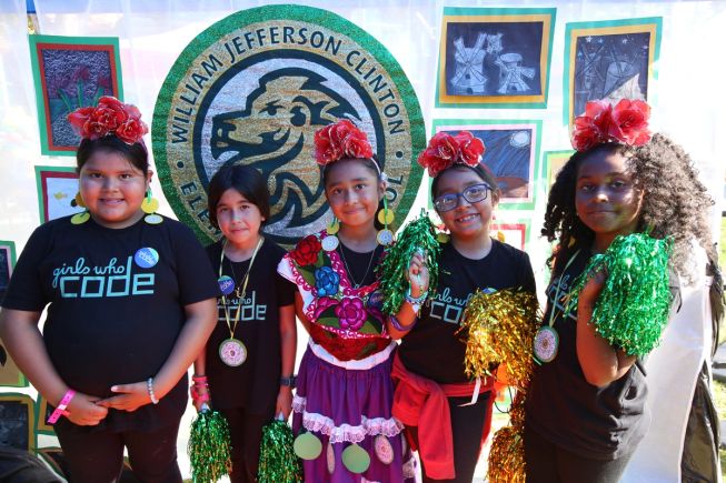 A group of young girls wearing “Girls Who Code” T-shirts standing together at a community event, smiling in front of a decorated display.