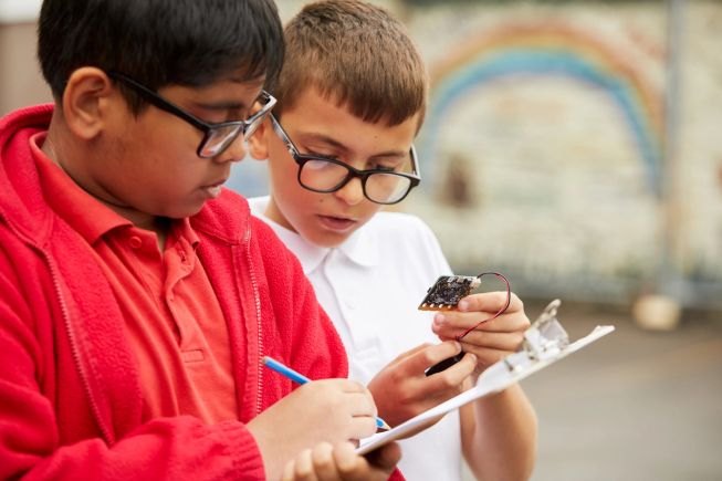 Two boys in primary school uniform standing in the playground. One is holding a with a clipboard and pencil making a note, and a the other is holding a micro:bit and battery pack, looking at the LED display.