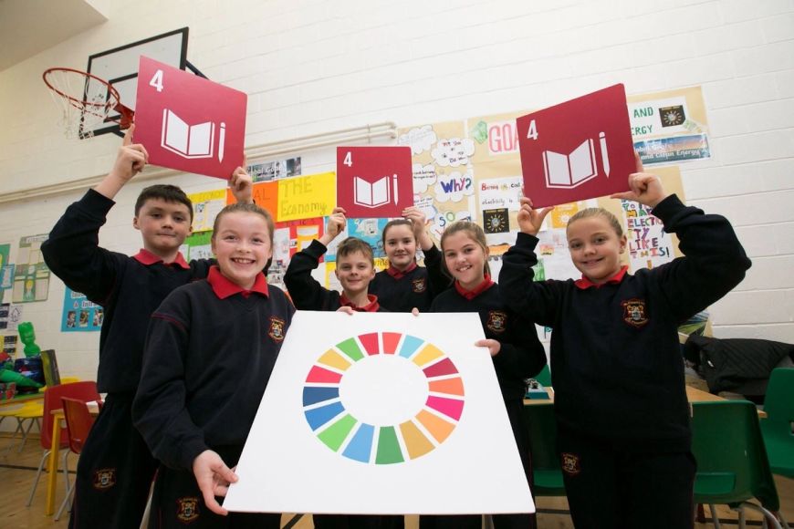 A group of primary/elementary age students stand in a school hall with a display board and basket ball hoop visible behind them. They are holding up the Global Goal 4 -Quality Education logo and the Global Goals wheel on large pieces of card.