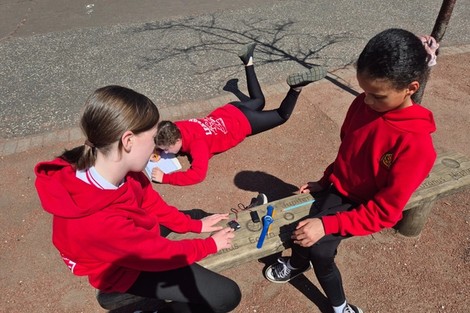 Three children sit outside as they measure temperatures of surfaces with the micro:bits