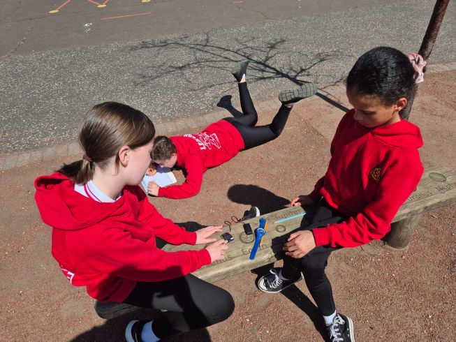 Two students sit on a wooden bench outside, with a micro:bit on the bench to measure the temperature. A third student is laying on the floor, recording notes on a clipboard