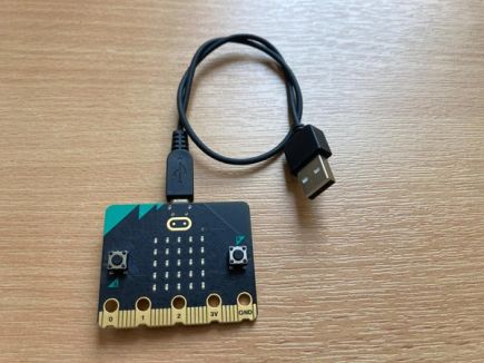 A micro:bit V2 lies flat on a desk with a micro USB cable already attached and looped round itself.