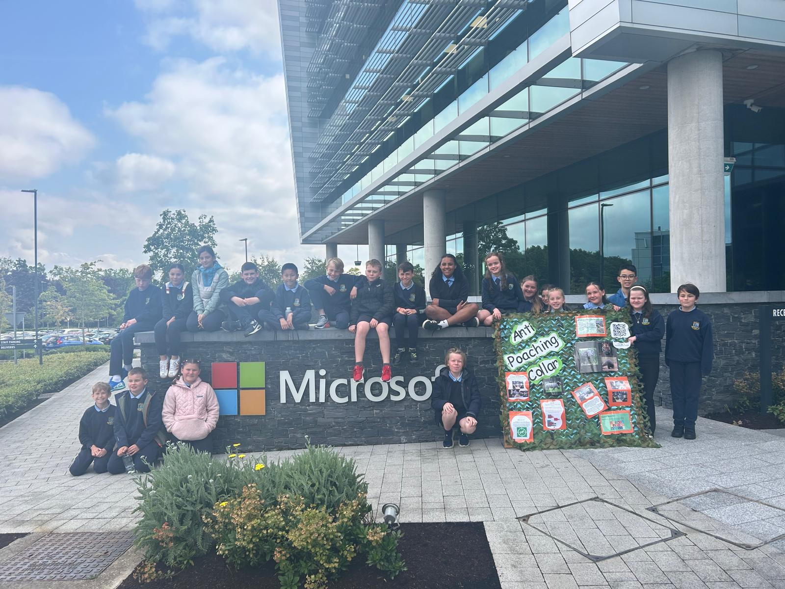 Students who worked on the project outdoors posing with their project poster next to a sign for Microsoft. 