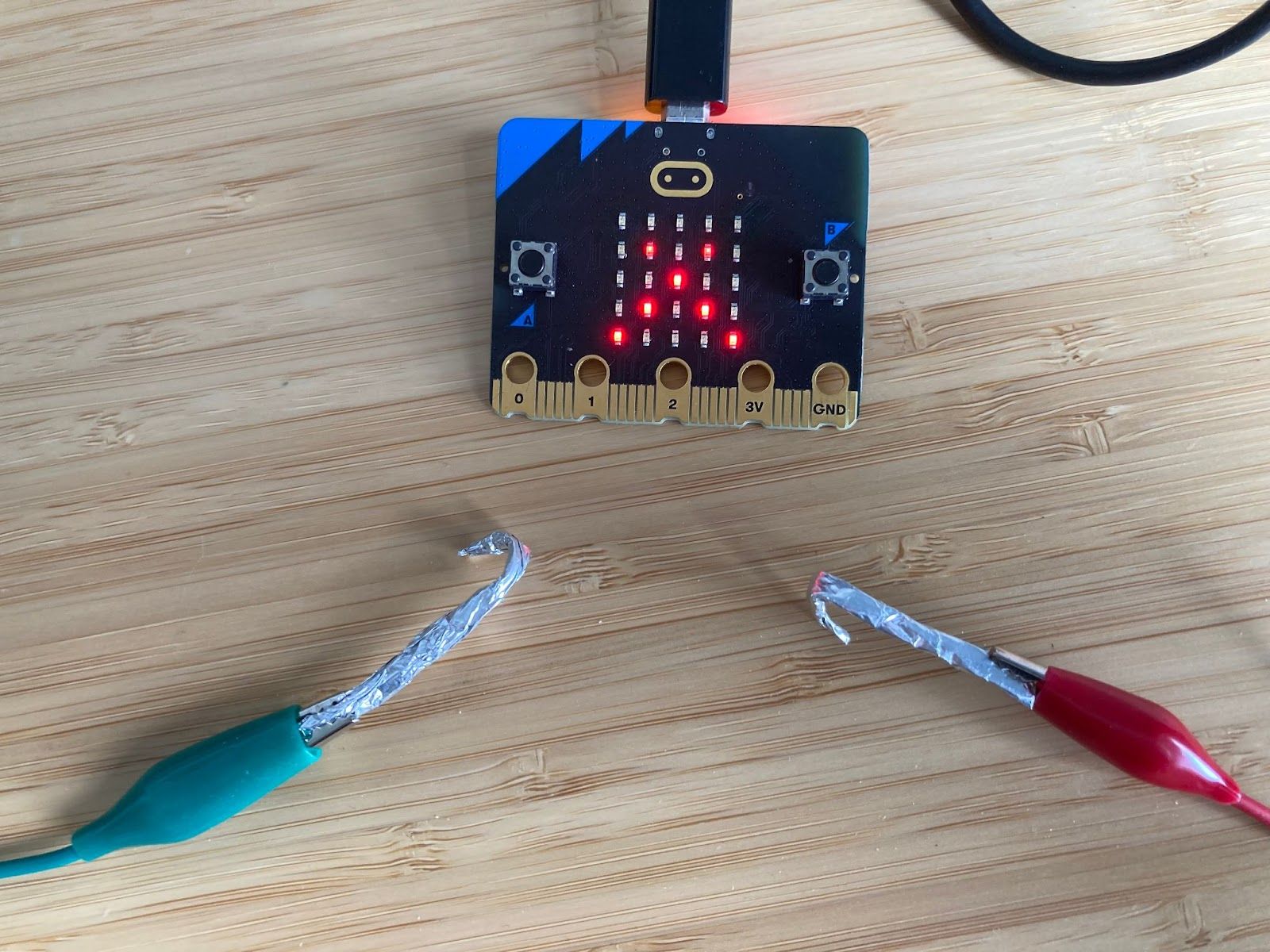 A micro:bit on a desk with green and red crocodile clips next to it. The crocodile clips are attached to hooks made of aluminium foil, ready to be connected to a micro:bit pin.
