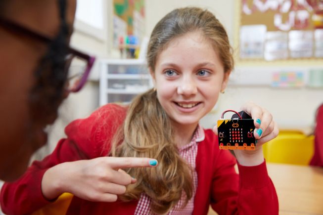 Merch mewn gwisg ysgol gynradd mewn ystafell ddosbarth, yn dal micro:bit gyda sgwâr bach ar yr sgrîn LEDS ac yn pwyntio ato.