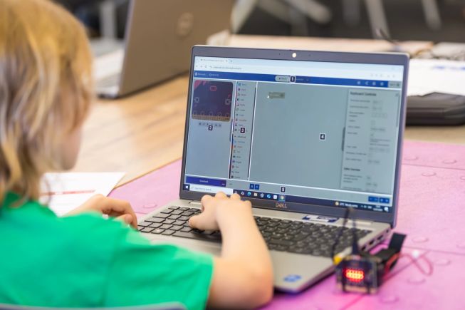 A child uses a keyboard to navigate Microsoft MakeCode on their laptop, with the micro:bit plugged in.