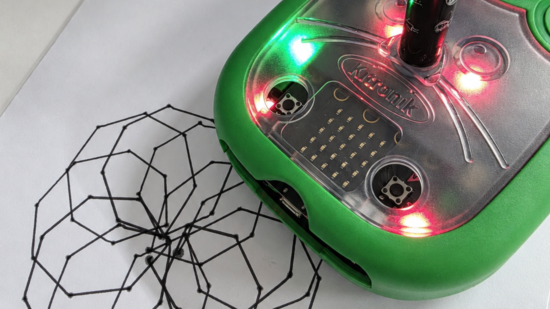 A green pen-drawing robot with illuminated LEDs sits beside the black geometric pattern it has created on paper.