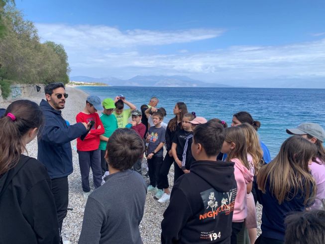 A male teacher stands on a beach, with a group of children stood around him listening to his explanation