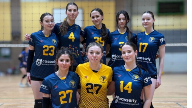 A girls’ volleyball team posing together on an indoor court in front of a net, wearing matching blue and yellow kits.