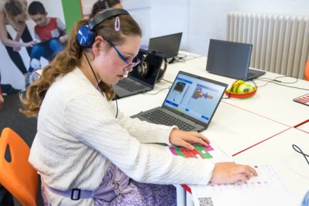 A student feeling the completed Shake alarm program with one hand and the tactile toolbox key with the other. The key shows tactile icons and braille labels for toolbox categories. They sit in front of a laptop with the MakeCode editor and corresponding project open.