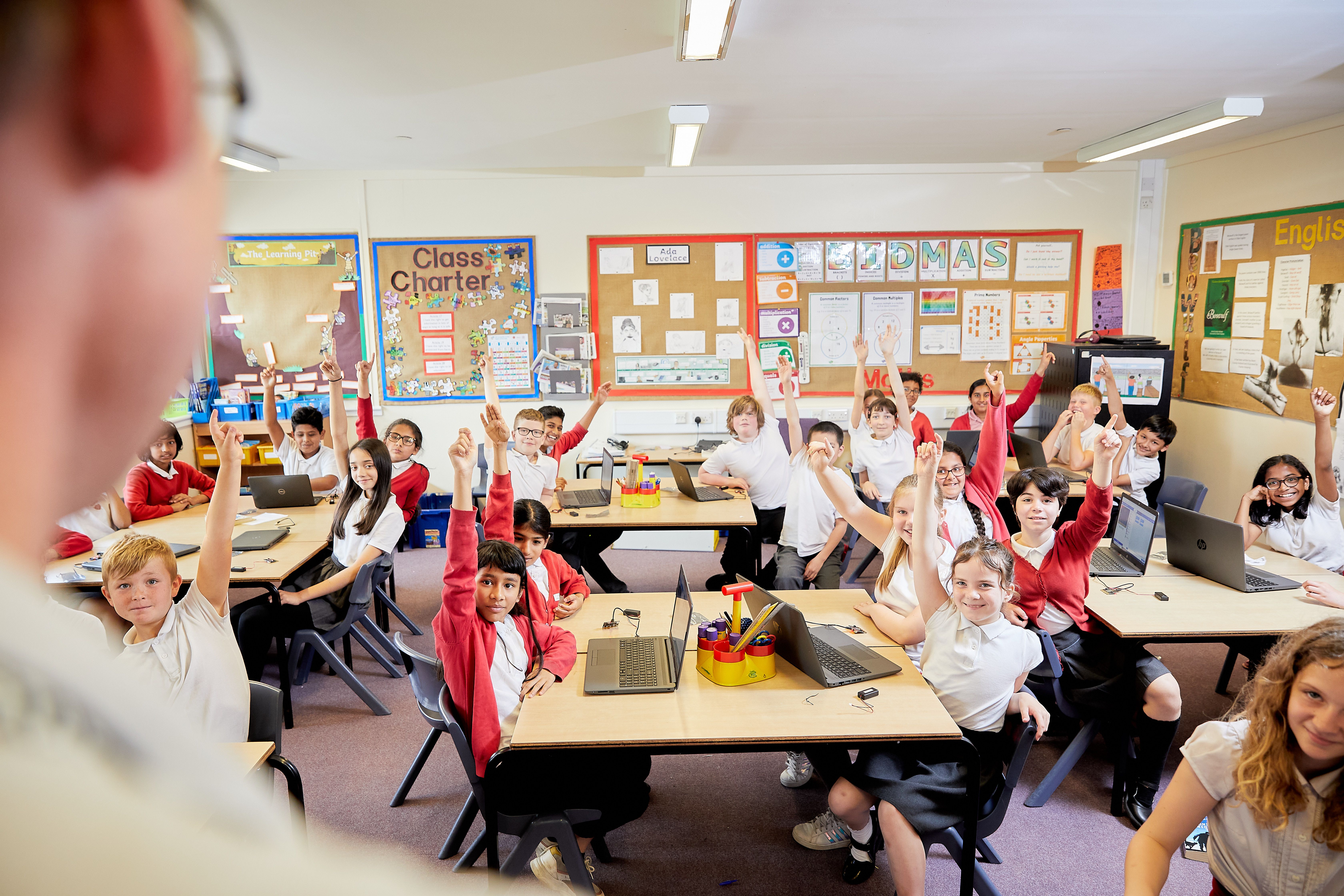 Seven children in school uniform, sitting in their classroom, with their hands raised
