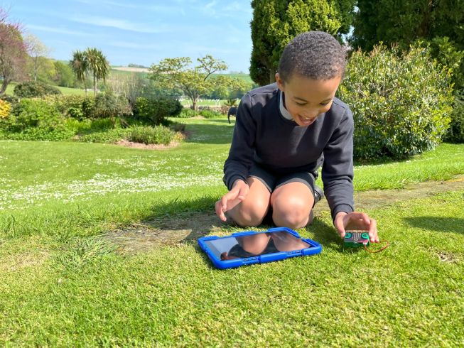 A boy at Windlesham House School kneels on the grass next to a tablet. He is holding a micro:bit excitedly