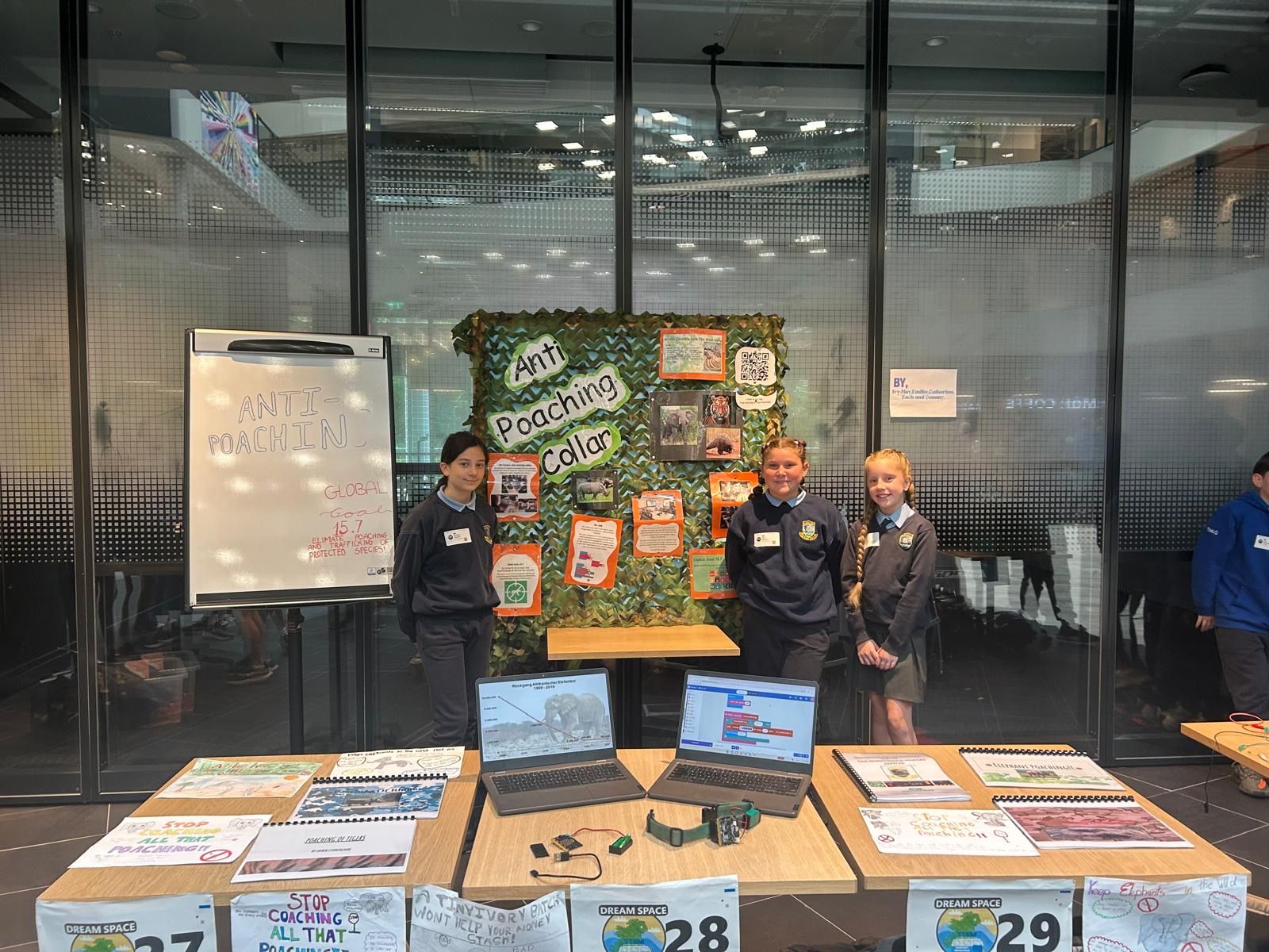 Two students post by the project display including two computers, a poster board, and several documents. 