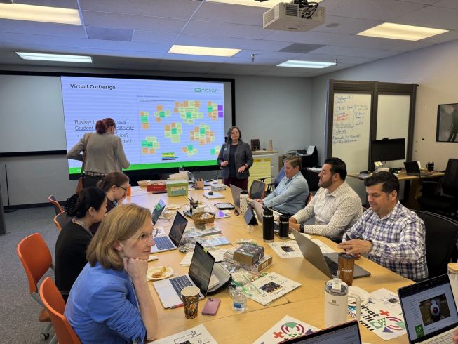 Educators and partners seated around a table with laptops and micro:bit kits, participating in a co-design session with a presentation displayed on a large screen.