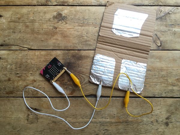 Sensor micro:bit with pin 0 connected to one foil pad, GND pin connected to another. A flap of cardboard with foil folds over and connects the two other foil pads when someone treads on it.