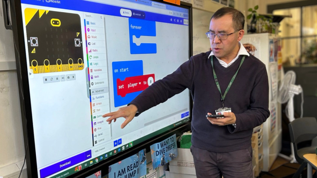 Allen Tsui points to a large screen displaying a micro:bit MakeCode project in his classroom.