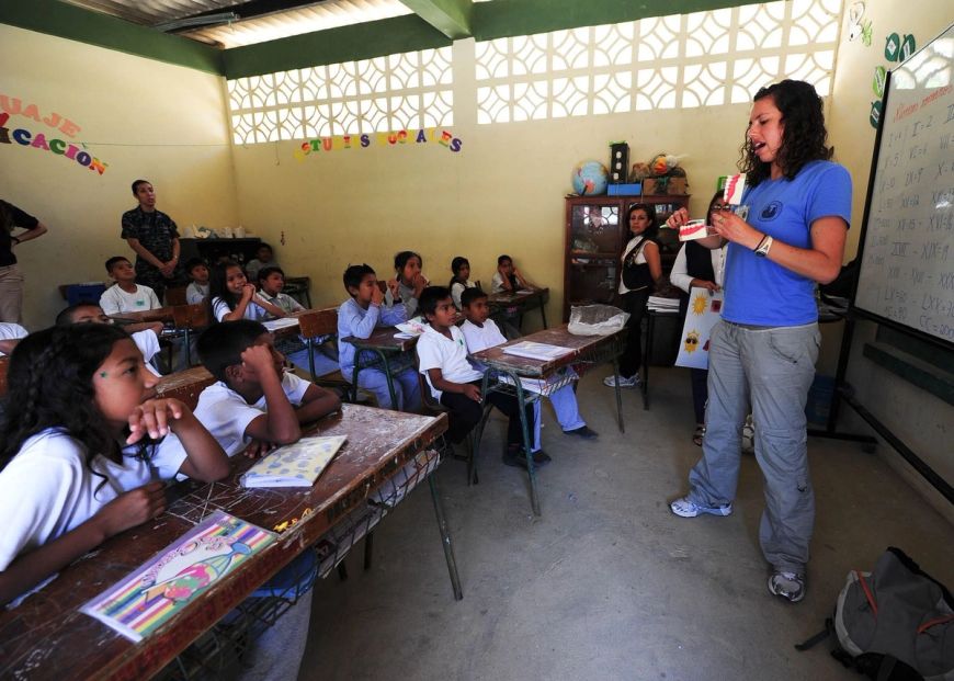 Imagen de una clase con una pizarra blanca al frente, pupitres de madera y equipamiento básico. Los niños escuchan a una maestra que sostiene unas imágenes de dientes y encías. 