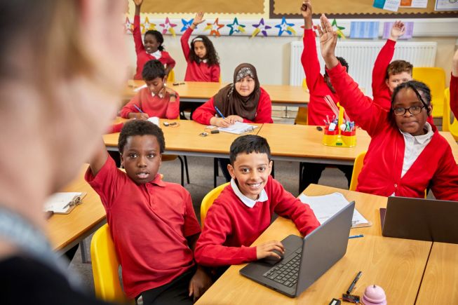 Image of 9 pupils in a classroom, many with their hand raised, teacher just out of view at the front of the room.