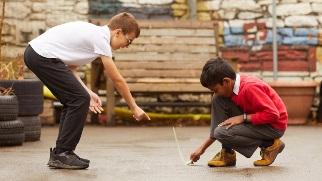 Two primary aged boys in school uniform in a playground are working together to draw a line with chalk on the ground. One is drawing and one is pointing where to draw.