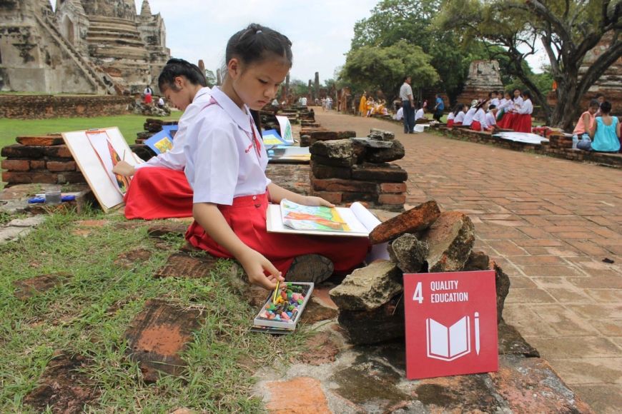 2 estudiants de primària/escola elemental asseguts a l'exterior amb un edifici antic o un monument al seu darrere. Hi ha dibuixos del seu entorn. Un objectiu global 4 - El logotip d'Educació de qualitat està sostingut al seu costat.