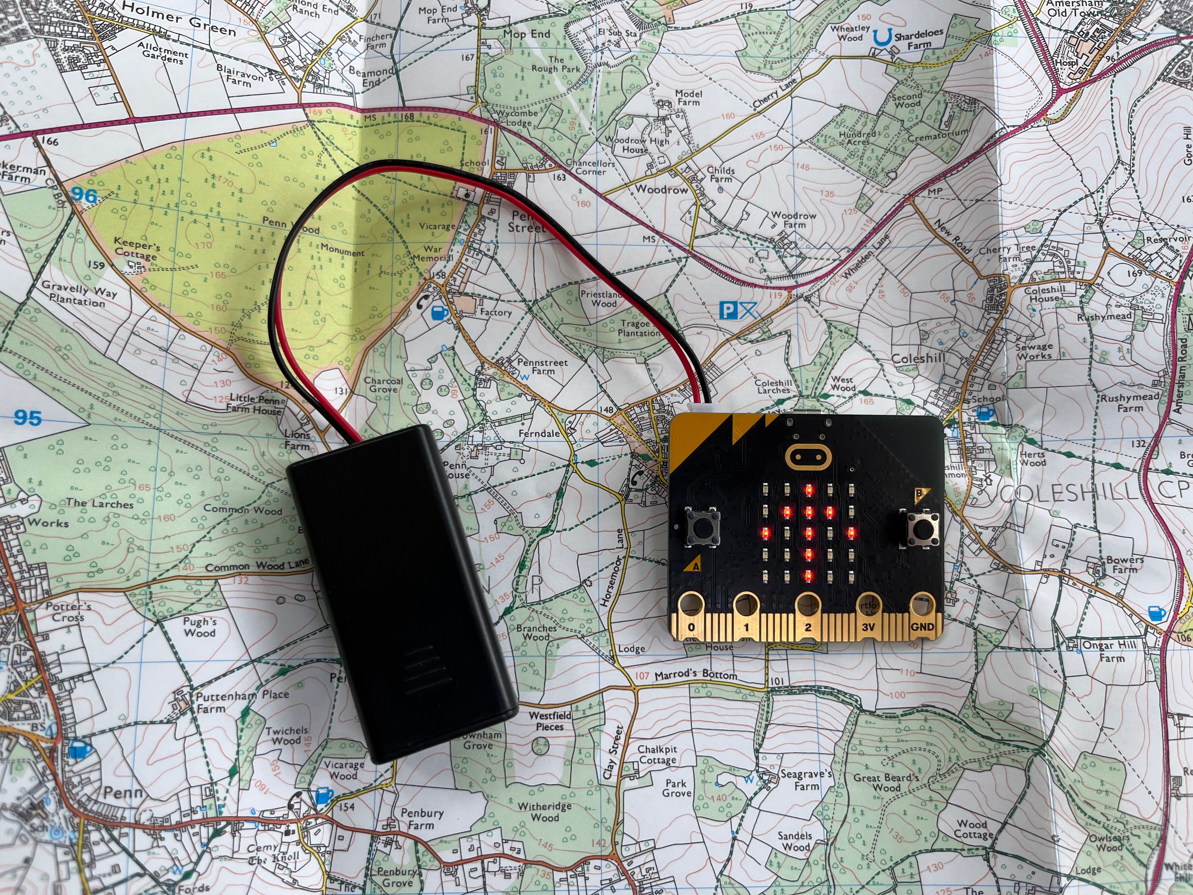 a micro:bit and attached battery pack resting on top of a map with an up arrow displayed on the micro:bit LED display 