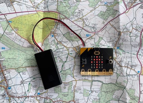 un micro:bit y una batería conectada descansando sobre un mapa con una flecha hacia arriba mostrada en la pantalla LED del micro:bit 