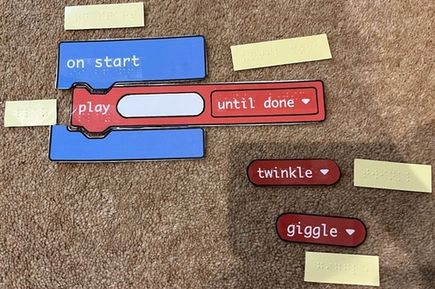 Large printed code blocks on card with braille labels have been placed on the floor to make a program that says on start; play (empty space) until done.