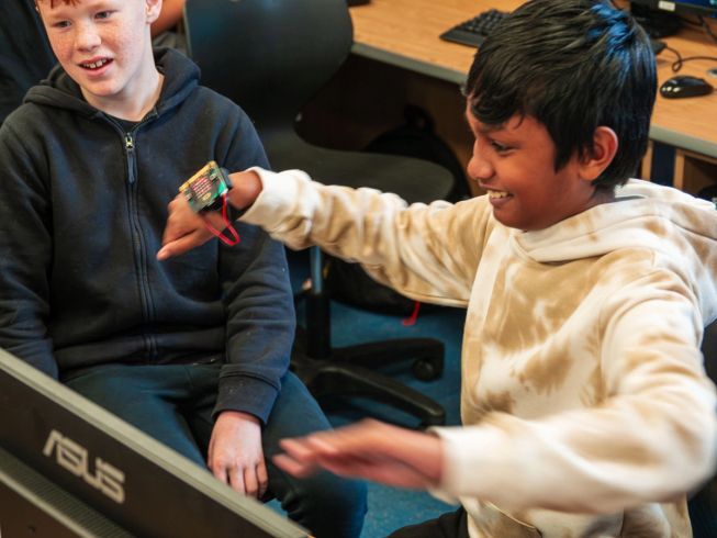 A student has a micro:bit attached to his wrist and is waving it around - another student looks on