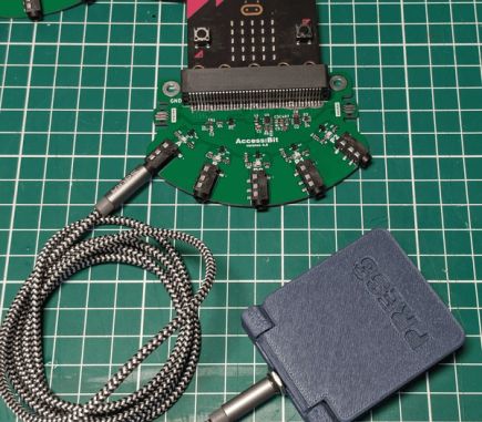 The Access:bit is a semicircular breakout board with a micro:bit slotted into an edge connector at the top edge, and five 3.5mm ports at the curved bottom edge. One port has a 3d-printed paddle switch attached.