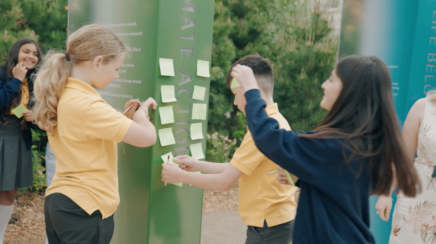 Children in school uniforms put sticky notes on a green wall