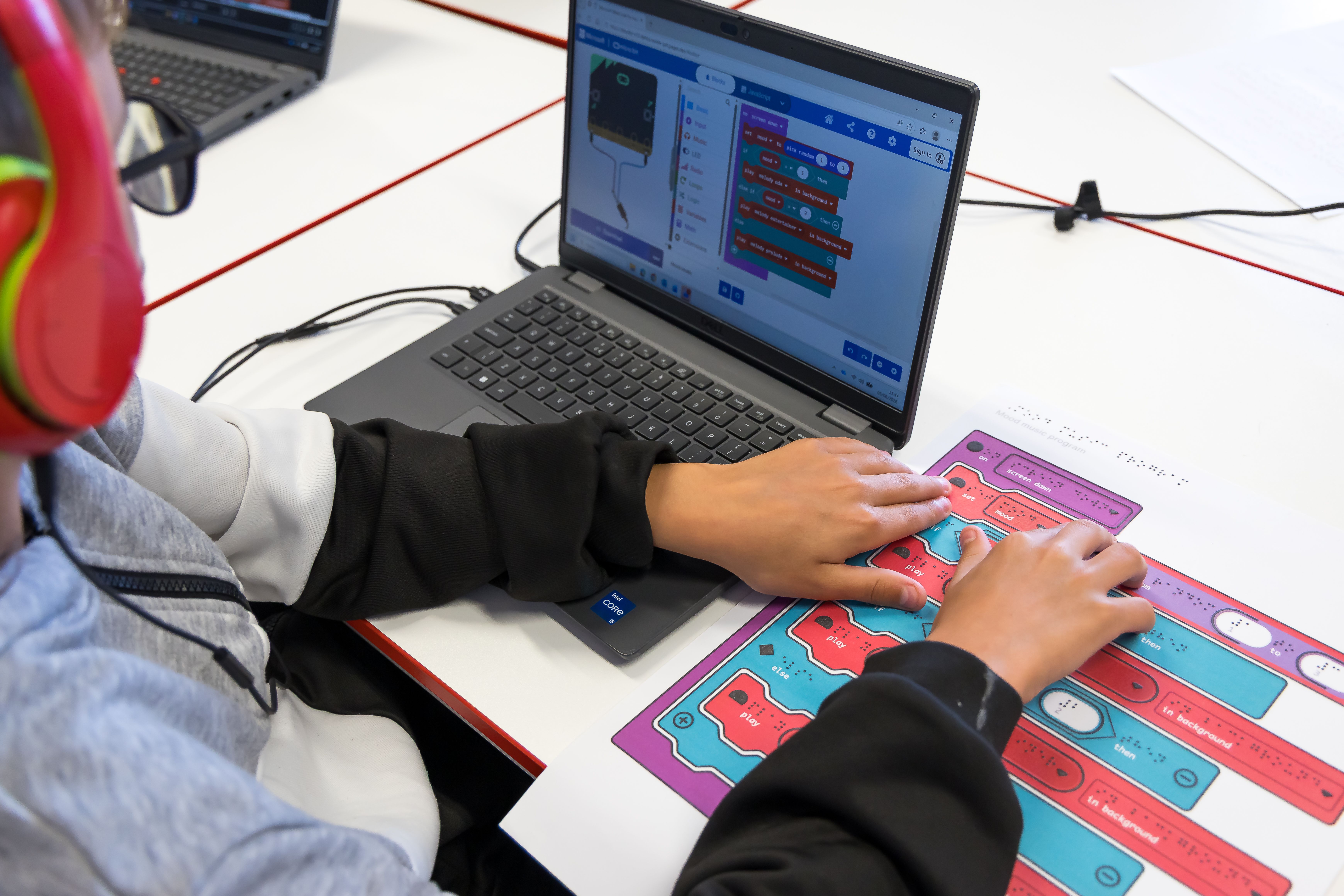 A student explores a tactile representation of a MakeCode program alongside the same block-based code displayed on a laptop.