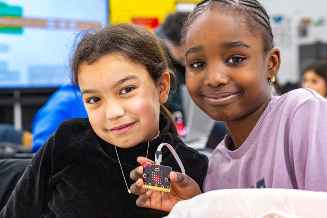 Three primary/elementary age students look at a micro:bit connected to a battery pack while outdoors. 