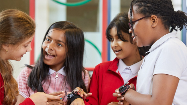 Mae pedair merch yn gwisgo gwisg ysgol gynradd, yn neuadd yr ysgol, mae dwy ferch â micro:bits ynghlwm wrth eu harddyrnau ac maent yn sgwrsio amdanynt.