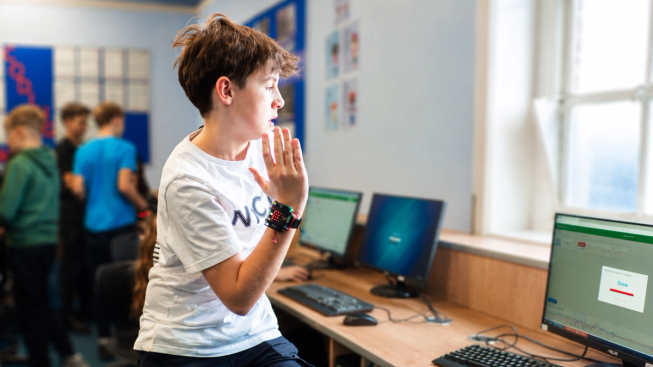 A young person wearing a BBC micro:bit on their wrist interacts with a computer by using hand movements in a classroom. Other students work together in the background, exploring physical computing activities.